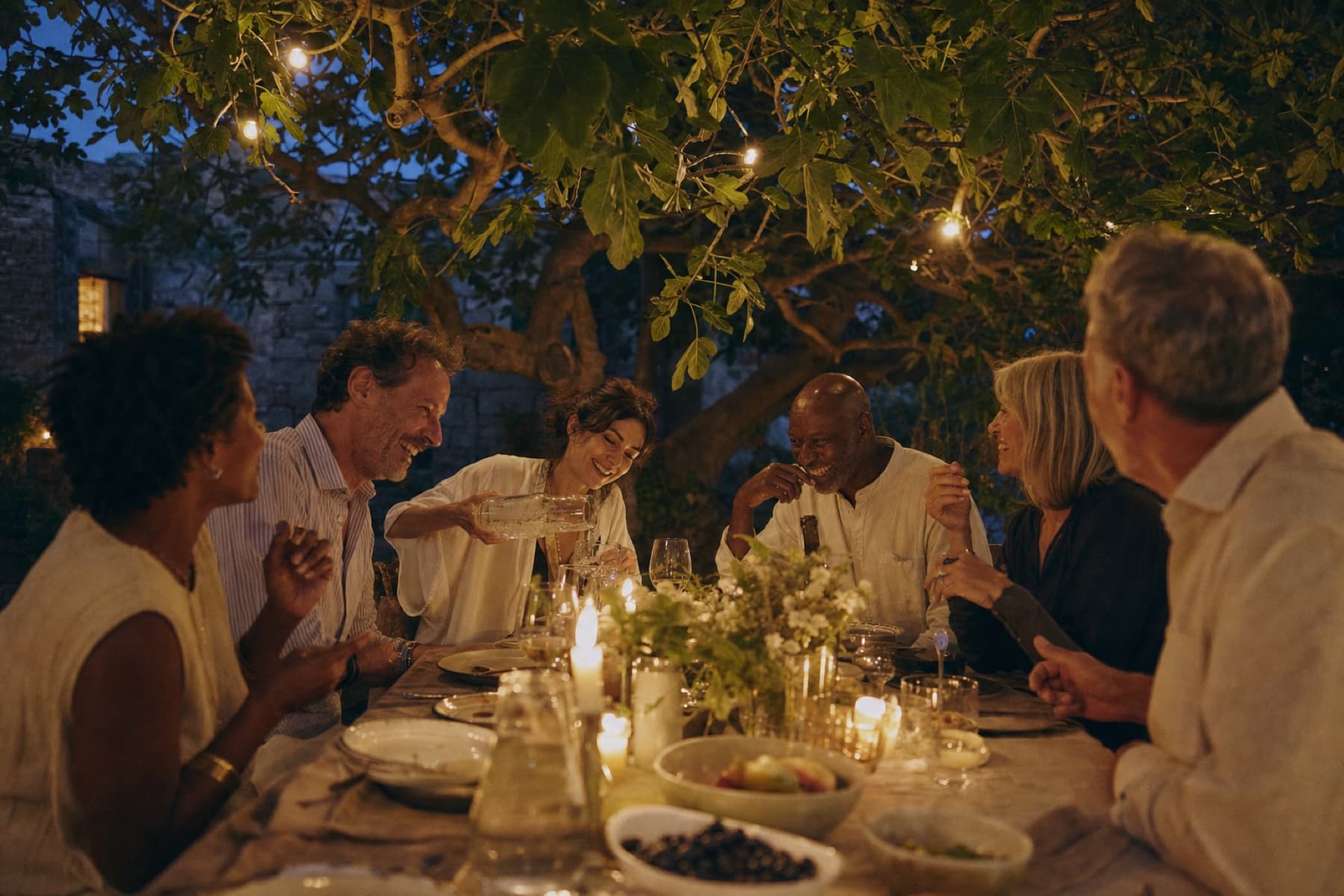 Six adults laughing at a long outdoor dinner table at dusk